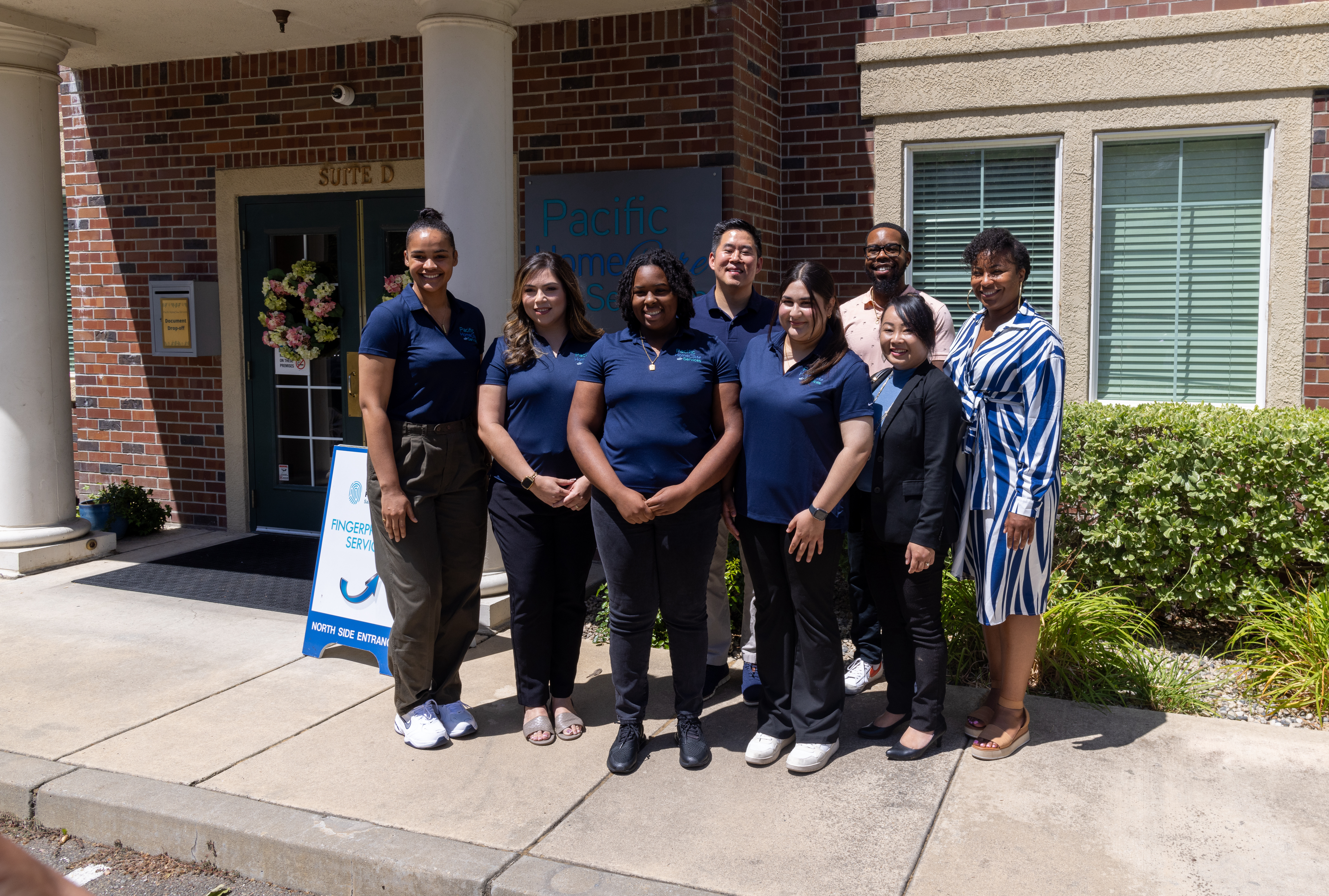 The image shows a group of eight professionals from Pacific Homecare Services posing together outside their office building. They are dressed in matching navy blue shirts, standing confidently in front of the building's entrance, which is marked by a sign that reads "Pacific Homecare Services." A welcoming wreath hangs on the door behind them, and a sign for fingerprint services is also visible, indicating the range of services offered by their office.