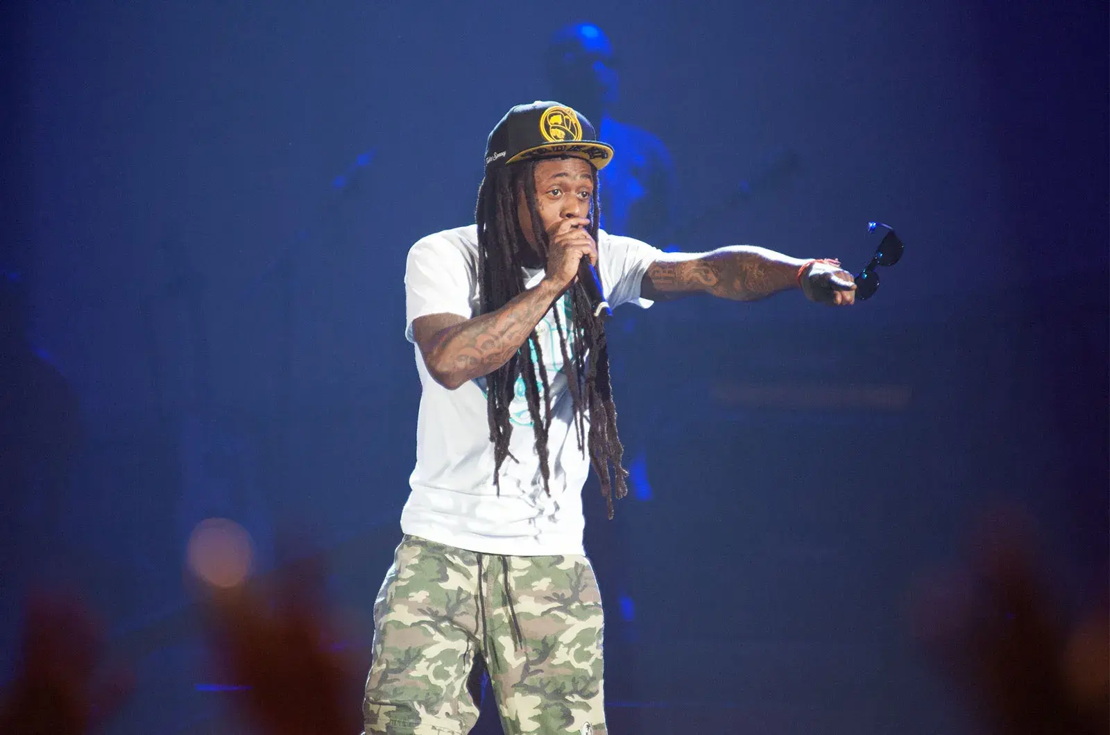 Lil Wayne performs at a live concert, showcasing his unique style in a white T-shirt and camouflage pants, complemented by a distinctive gold cap. Engaged with the audience, he delivers his lyrics with intensity under the bright stage lights, exemplifying the dynamic energy he is known for.