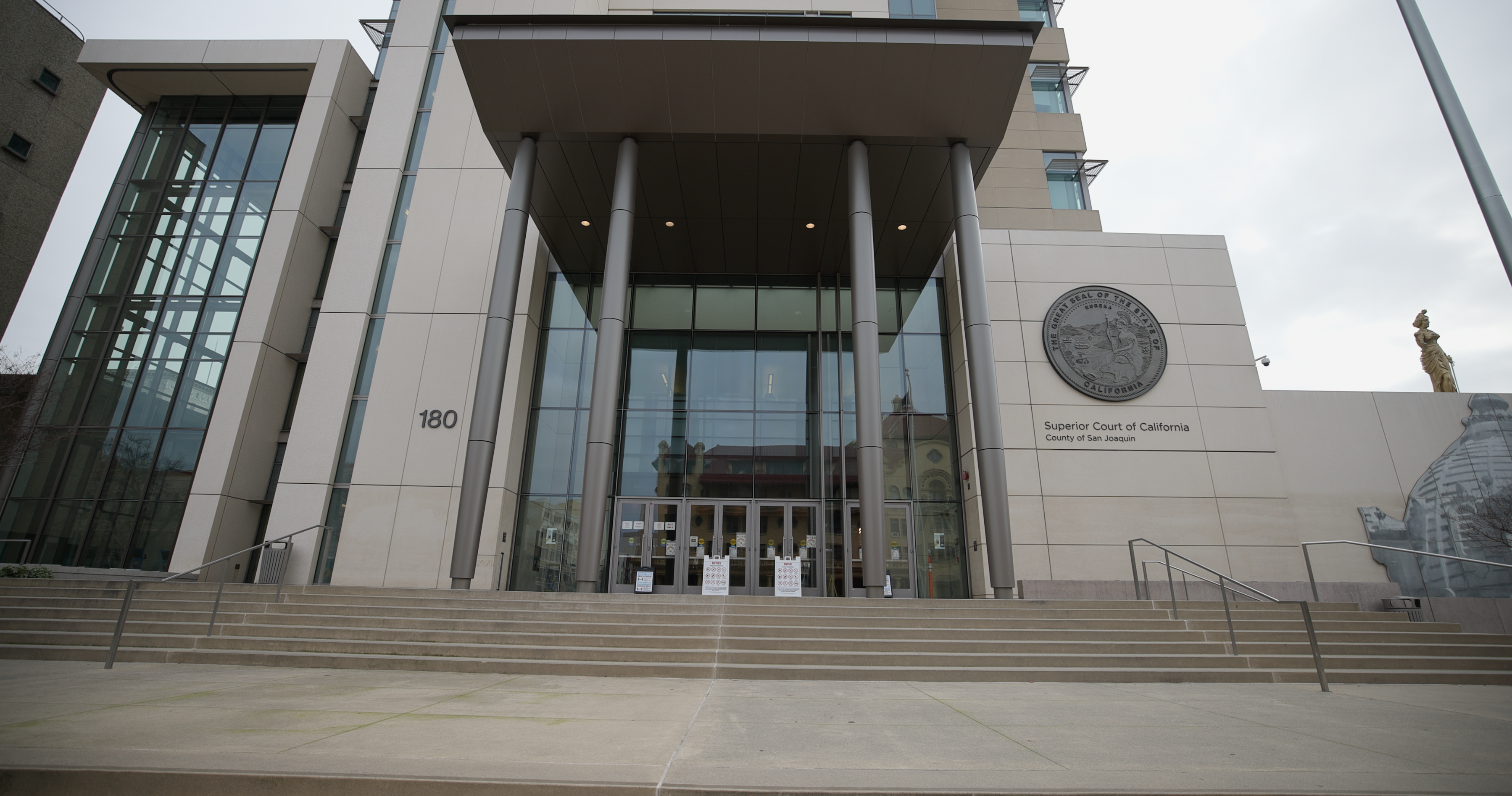  San Joaquin County Superior Court in downtown Stockton, California. The building features a large glass entrance framed by metal columns, with the court's seal prominently displayed on the wall to the right. Stairs lead up to the main doors from the street level.
