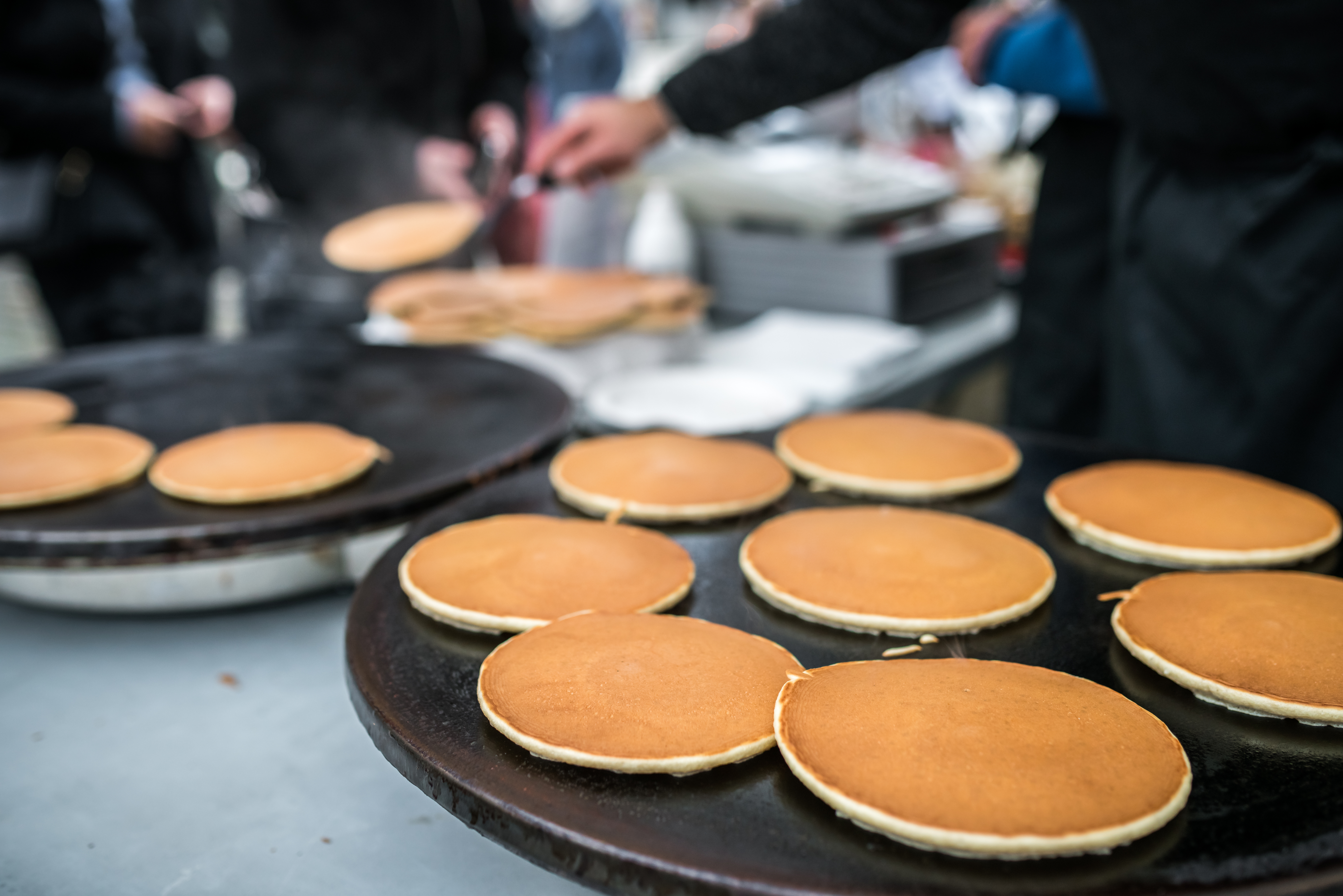 Image of freshly cooked pancakes on a large griddle, being prepared at an outdoor community event, with blurred figures of people serving in the background.